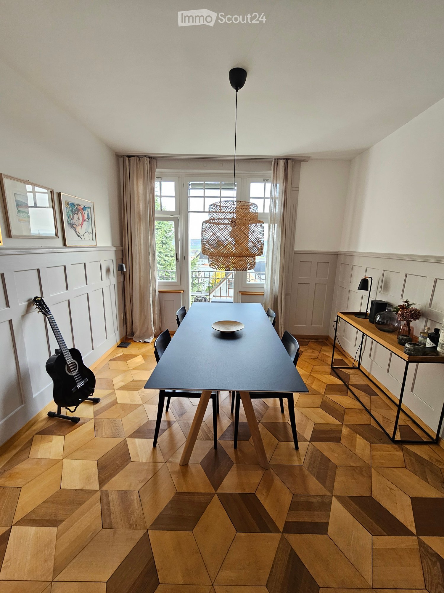 Geometric wooden floor, long table with six chairs, modern pendant light, guitar leaning against the wall, two large windows, large lamps