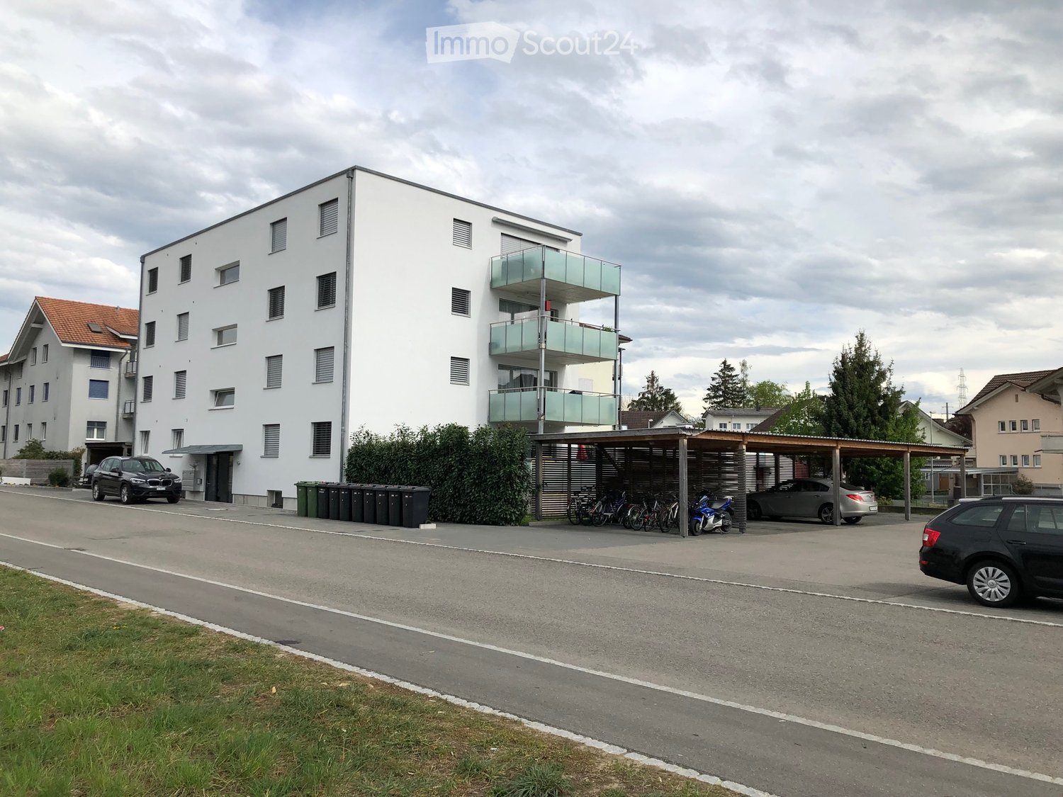A four-story building with white walls, balconies on upper floors, car parked in front, bikes under a canopy