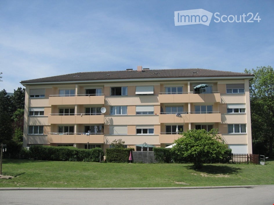 Three-story apartment building, beige exterior, multiple windows, balconies on every floor, well-maintained garden, street view