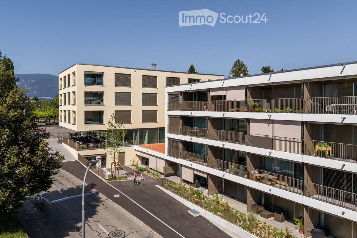 Modern architecture, beige color, several floors, multiple balconies with railings, outdoor seating on some balconies, trees, mountains in the background, paved road in front.
