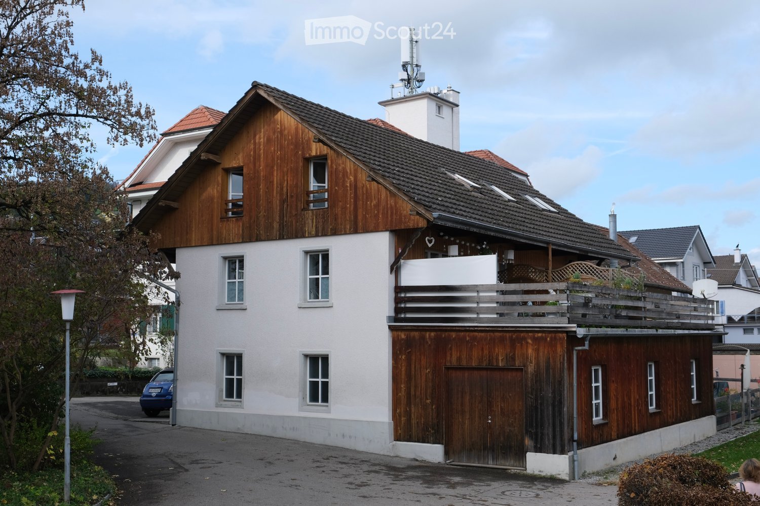 detached house, white and brown wooden exterior, multiple windows, brown tiled roof, balcony with wooden railing, car parked in front, greenery in front