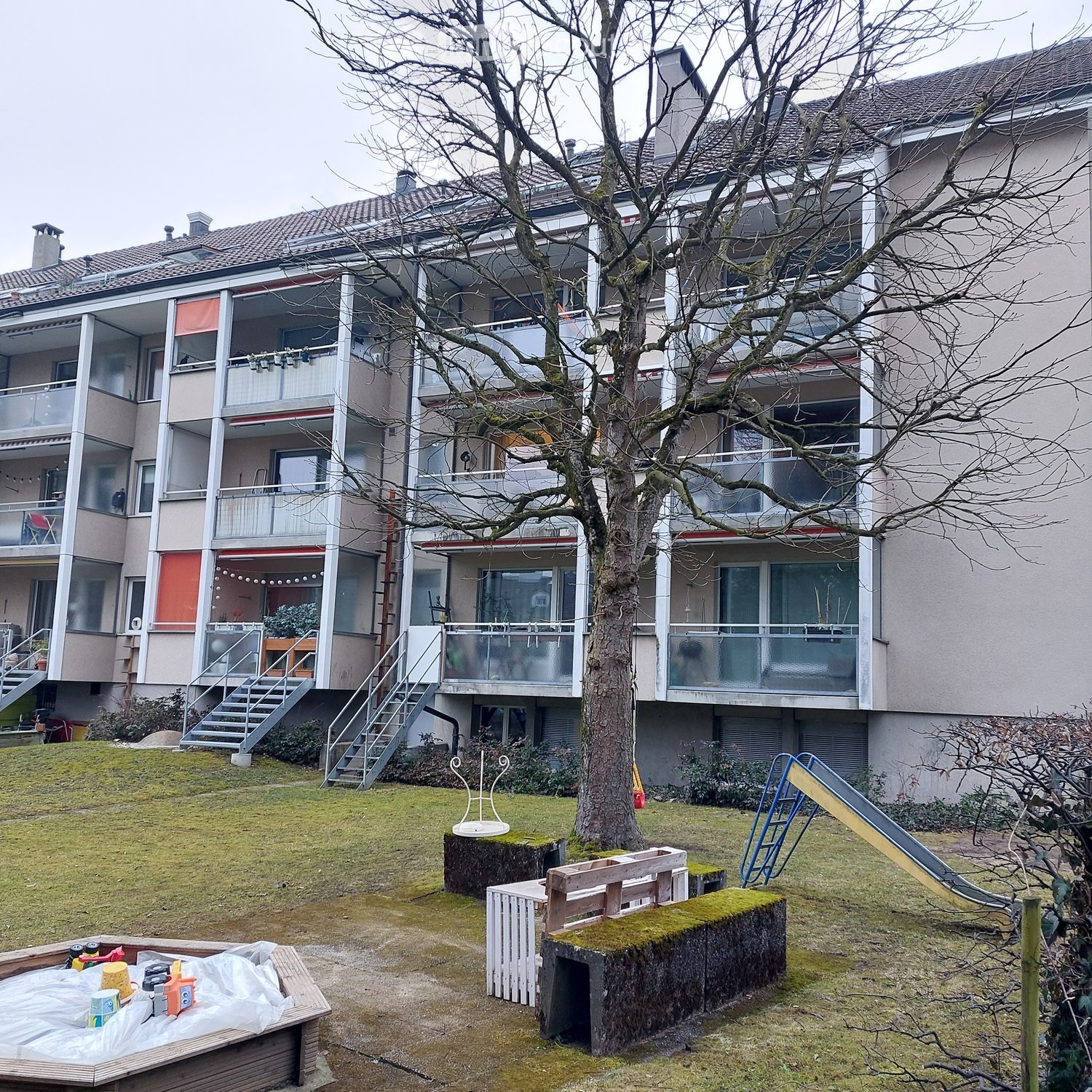 Apartment building with many balconies, tree in the middle of the yard, slide and benches in the yard