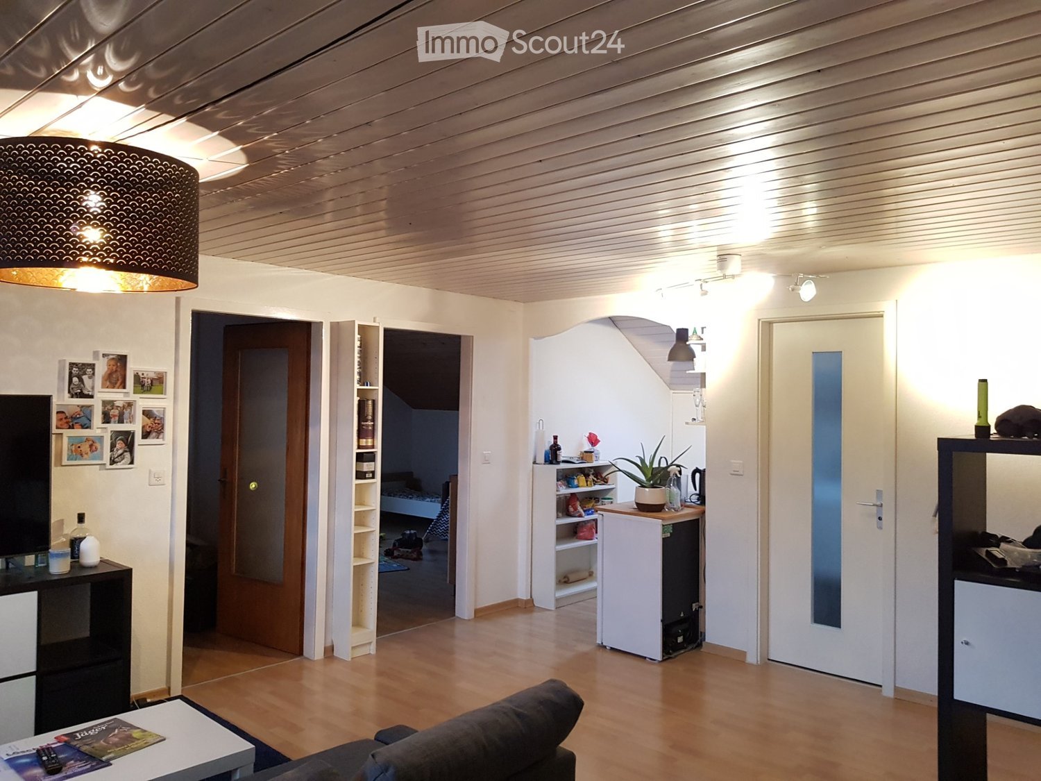 Wooden floor, ceiling with recessed lighting, living room with couch and coffee table, white walls, shelves with various items, a white fridge, TV, framed pictures on the wall, bedroom in the background.