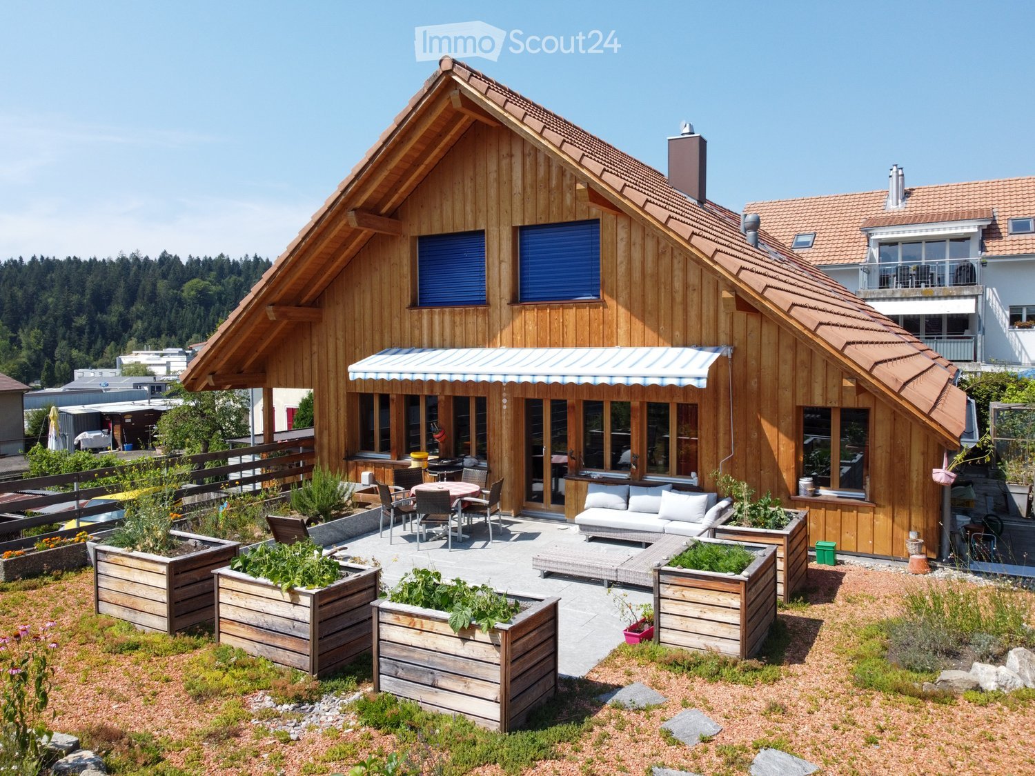 A wooden chalet house, brown roof, glass windows, terrace with wooden planter boxes, awning, and a garden with flowers