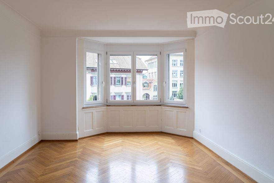Empty room with parquet flooring, bay window, white walls, three windows.
