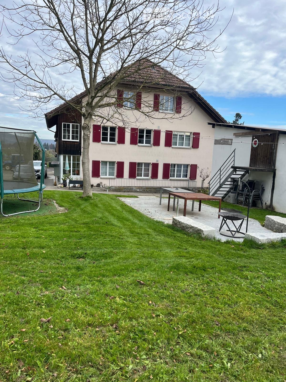 2 story house, white exterior, red shutters, outdoor table, seating area, trampoline, lawn, garage, stairs, car parked