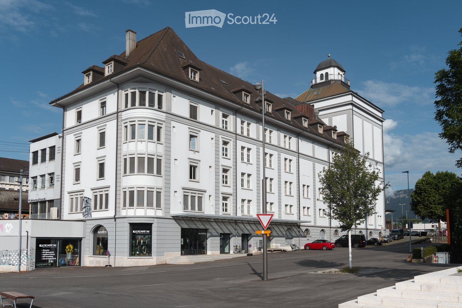 Multi-story building, white exterior, brown roof, many windows, shops on the ground floor, parking lot in front