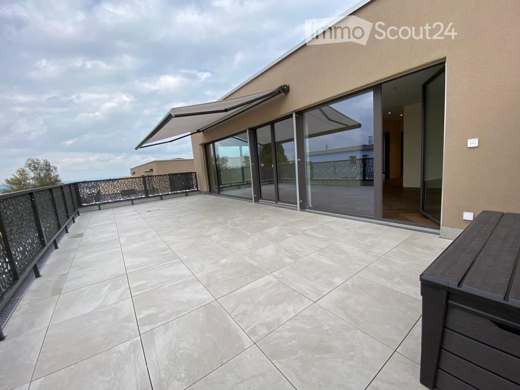 large balcony with tiled flooring, retractable awning, glass sliding doors, black railings, distant view of trees and buildings