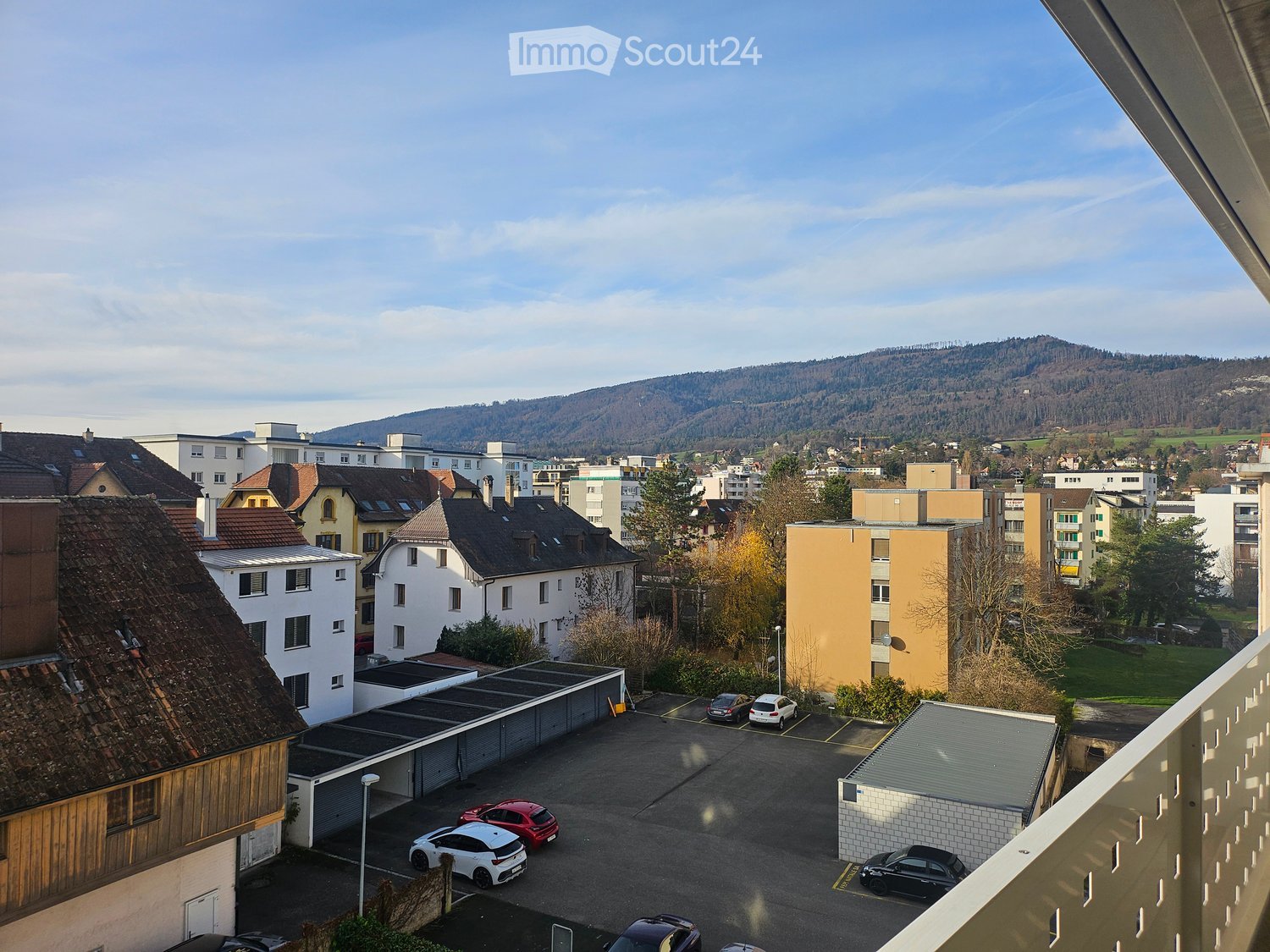 Town, multiple buildings, several parked cars, distant mountains, green trees, balcony, wide view