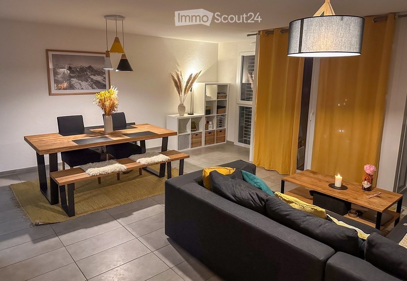 Living room with wooden table, dining chairs, a rug, a couch, coffee table, two lamps, a window, shelves, picture frame, and a floor-mounted lamp.