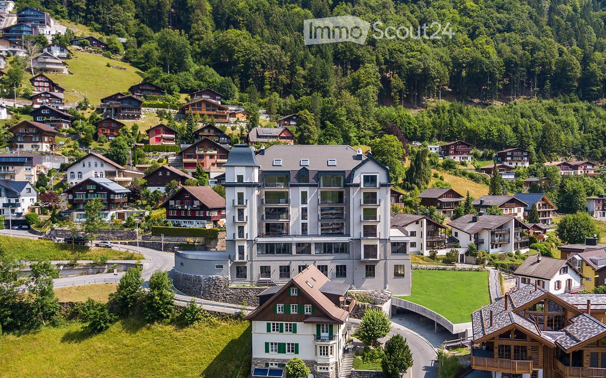 Aerial view of a modern apartment building surrounded by houses and trees, set on a hillside with a scenic valley below.