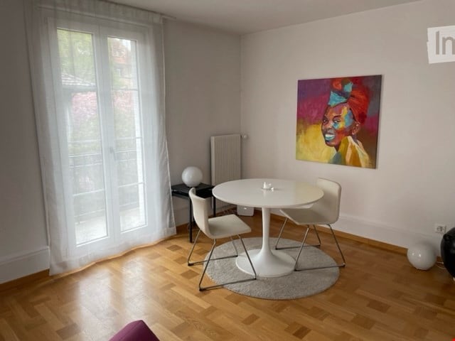 Dining area with a round white table, four white chairs, large windows with white curtains, a radiator, a small table with a white lamp, and a colorful painting