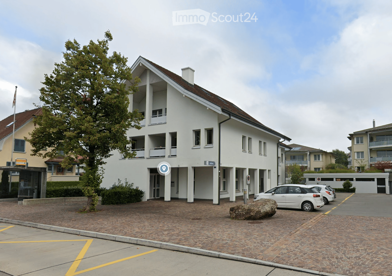 White multi-storey building, brown roof, windows, parking lot with parked cars, cobblestone pavement, tree, bushes, bus stop