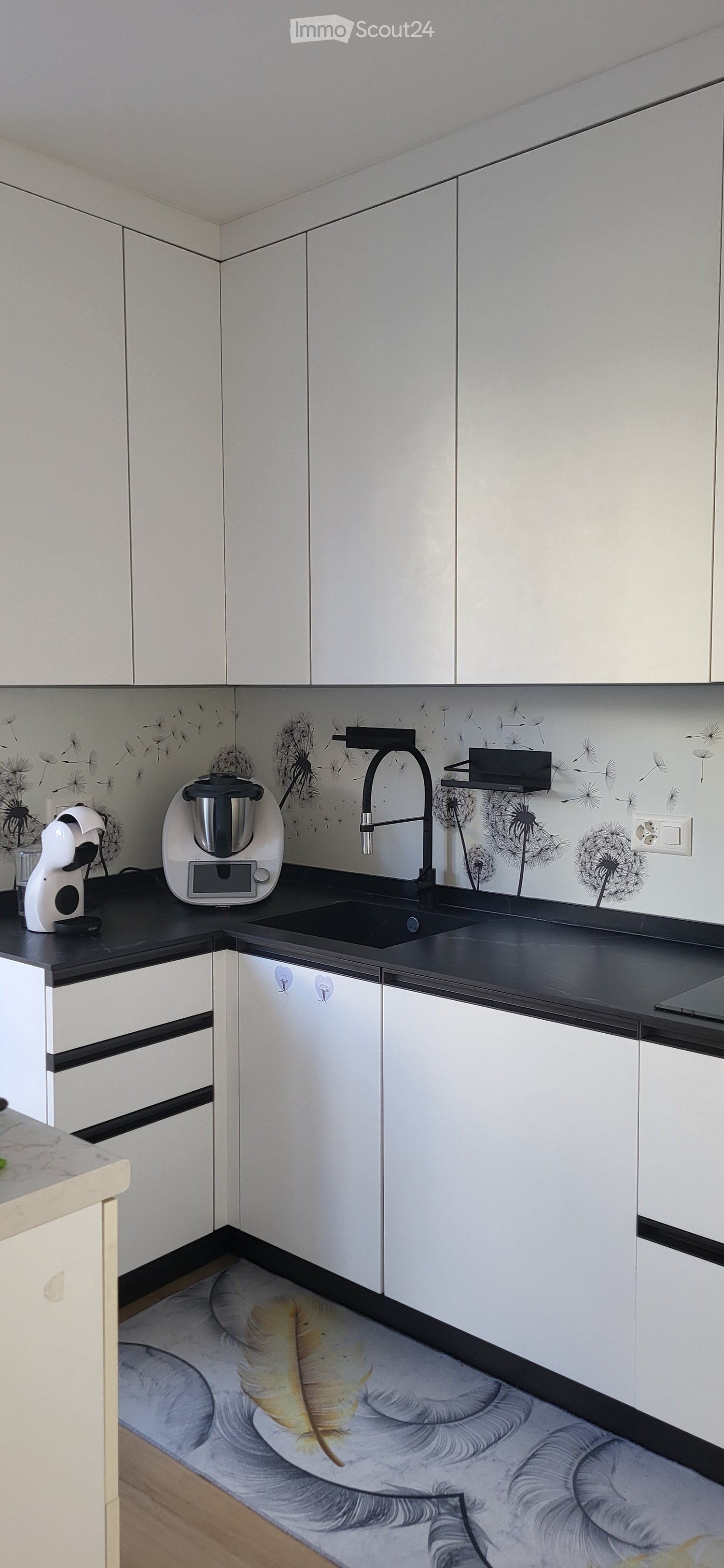 modern kitchen with white cabinets, black countertop, integrated dishwasher, washing machine, faucet, wall with dandelion print, coffee machine