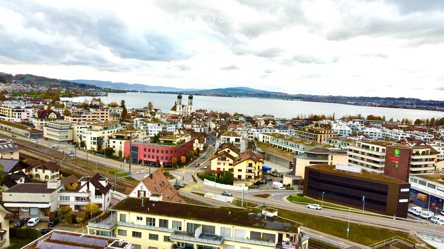 The image captures a panoramic aerial view of a cityscape by a lake with a cloudy sky. It shows various buildings of different architectural styles, including residential apartments, commercial structures, and green rooftops. The area is well-planned with