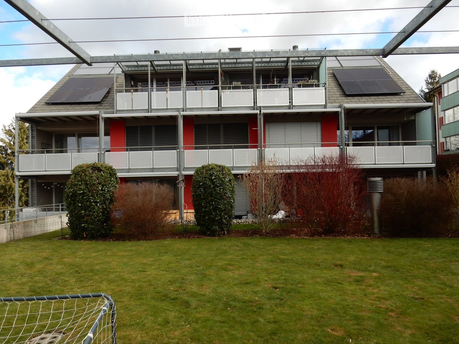 Multistory building, red exterior, metal railings on balconies, solar panels on the roof, raised ground floor
