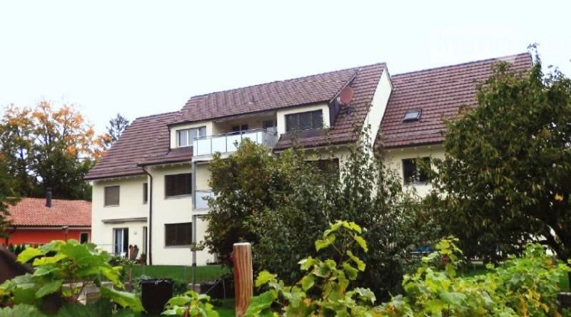 Two-story white house with brown roof, balcony on the second floor, garden with plants and trees