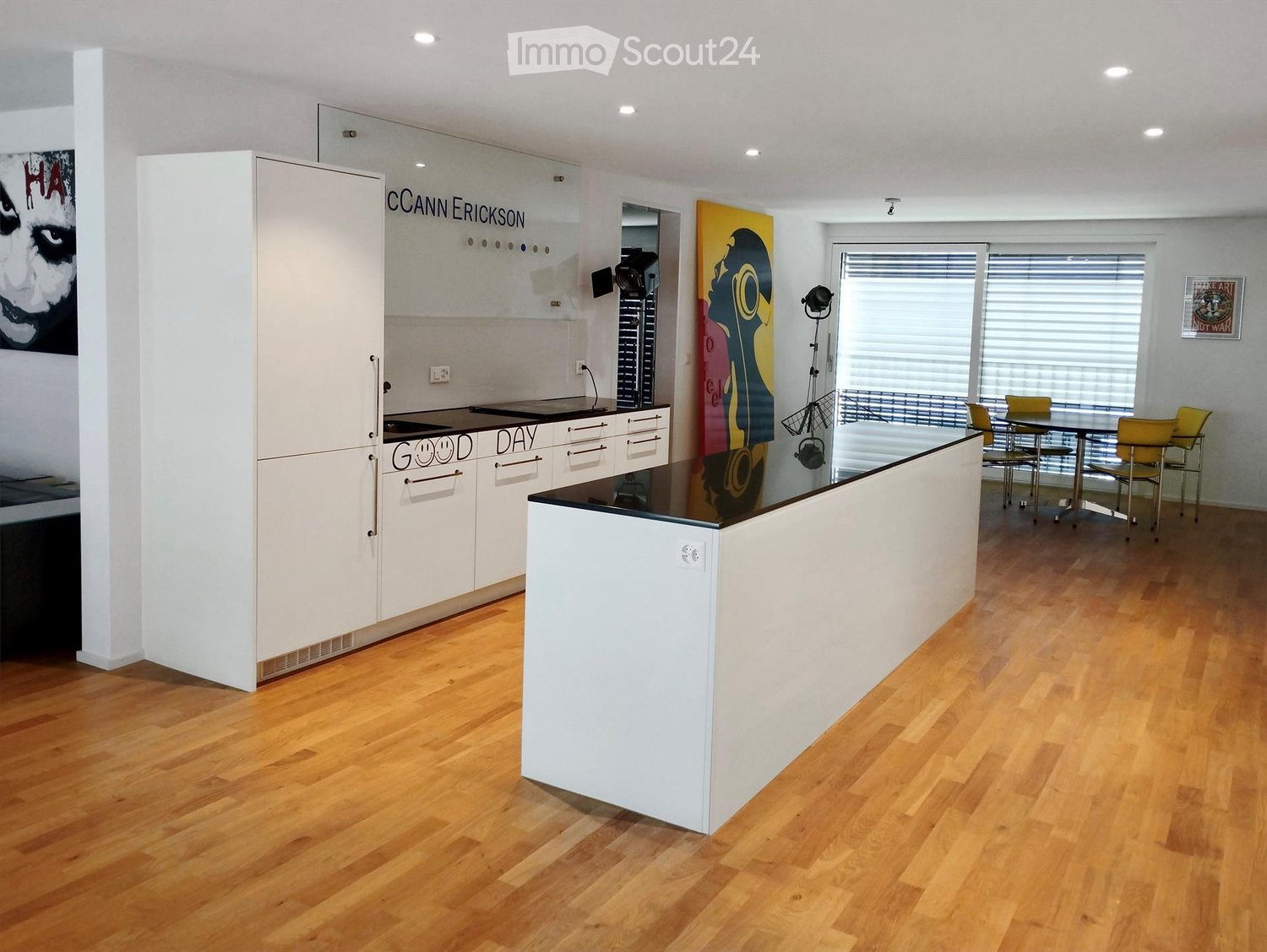 modern kitchen with center island, white cabinets, black countertops, dining area with a table and chairs, white walls, wooden floor, large windows with blinds, artwork on the wall, and a picture frame