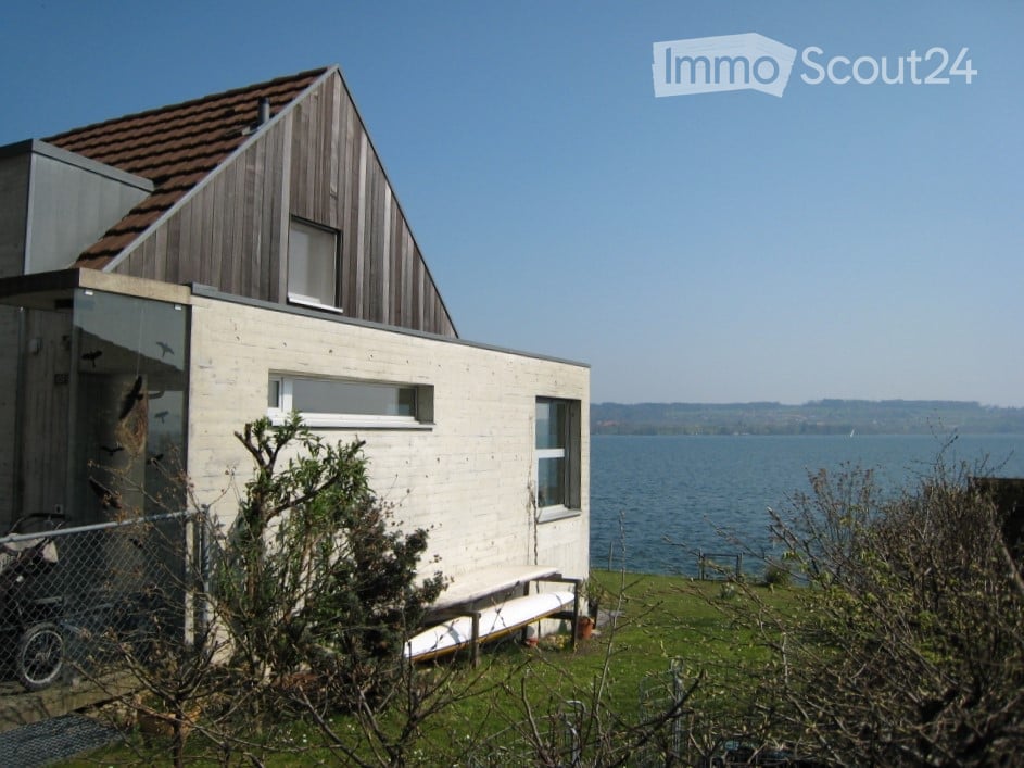 A modern house with a red tiled roof, a glass wall, a white bench in front, a large water body nearby, and a green field.