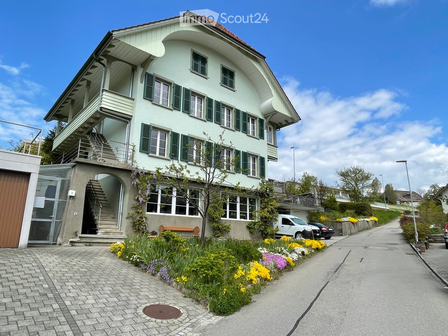 3 story building, raised ground floor, brown wooden door, window shutters, wooden bench, front garden with flowers, paved driveway