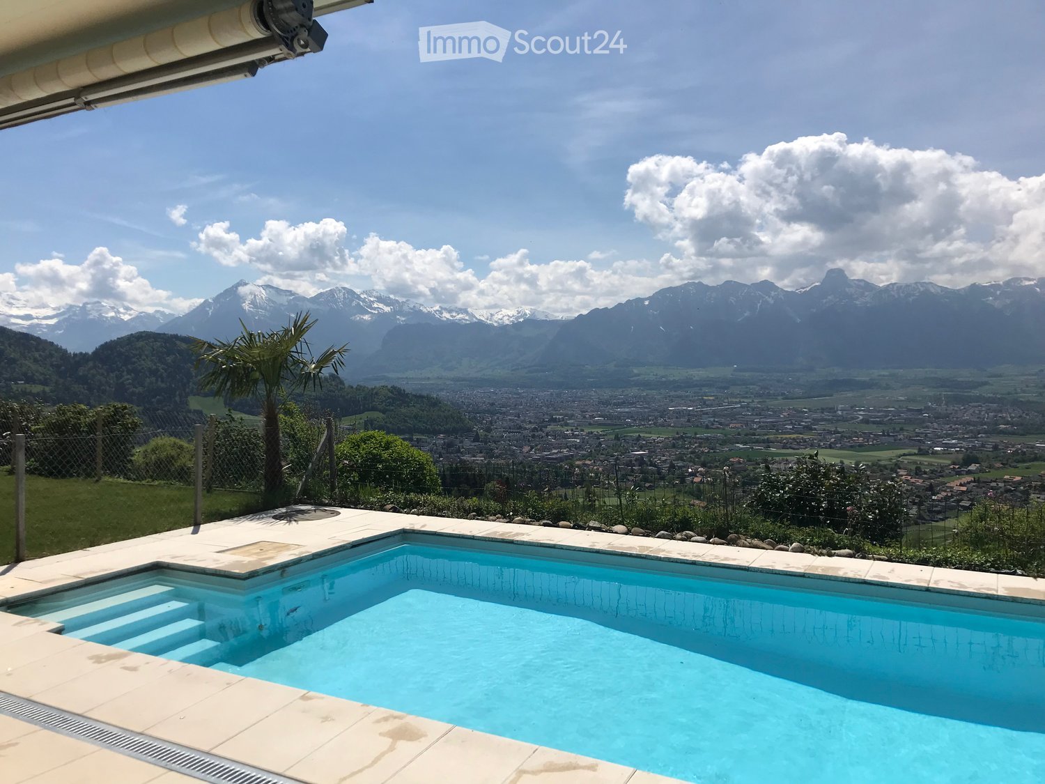 Swimming pool with steps, fenced garden with green grass, palm tree, mountain and town view