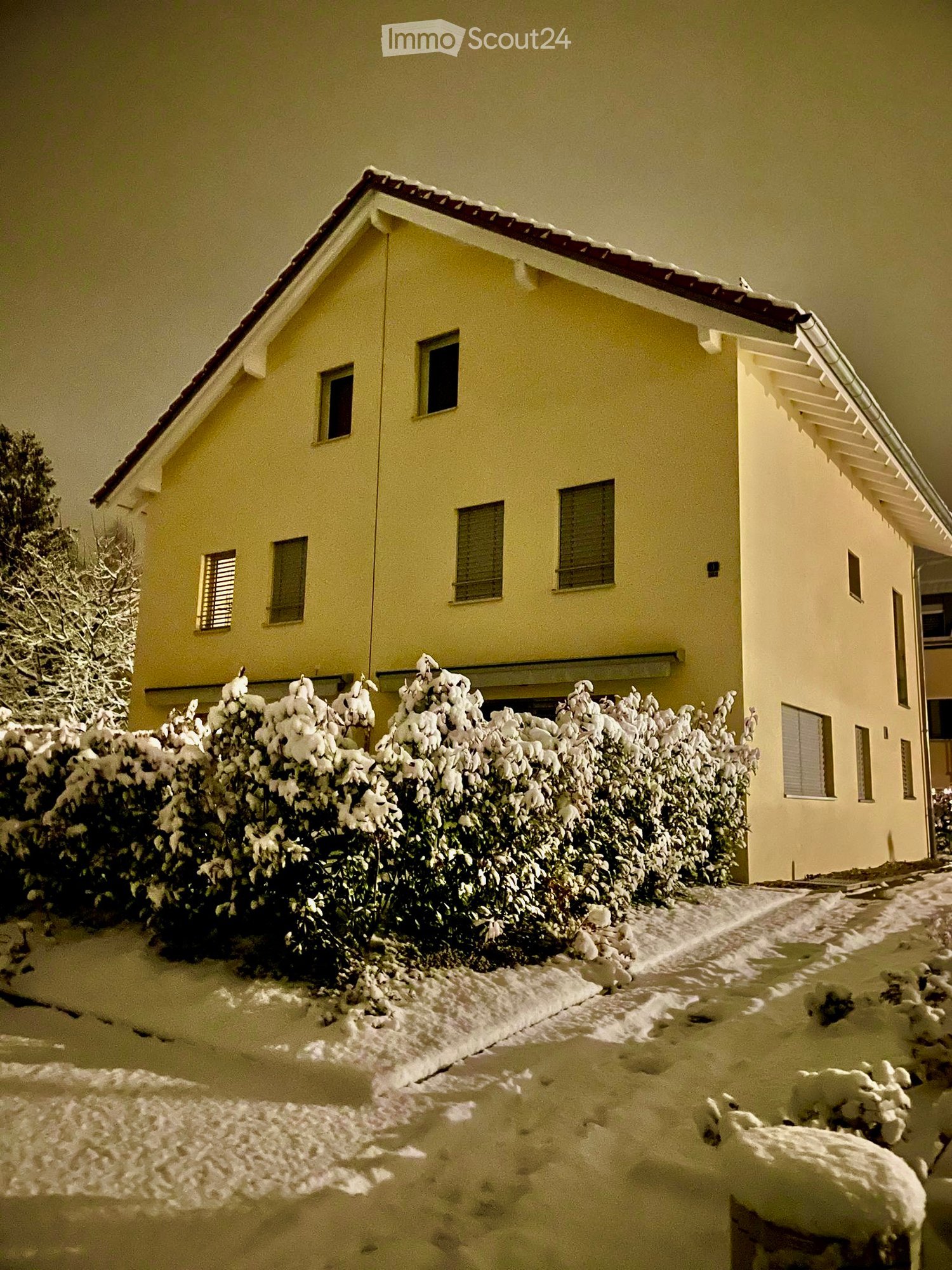 Two-story house with yellow exterior, several windows, snow-covered bushes, raised ground floor.