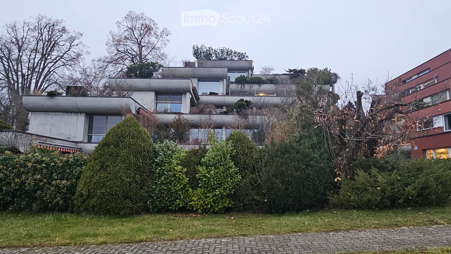 modern apartment building, grey facade, greenery on rooftop, multiple windows, shrubs and trees surrounding