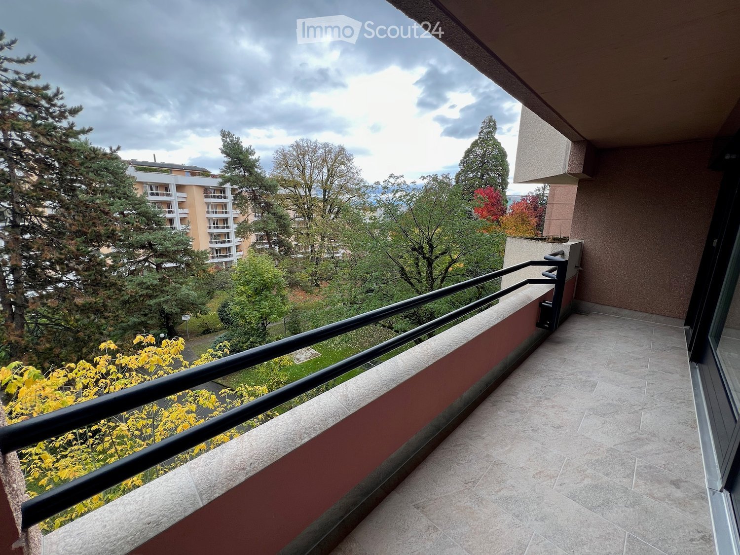empty balcony with tiled floor and black railing, trees and buildings in the background