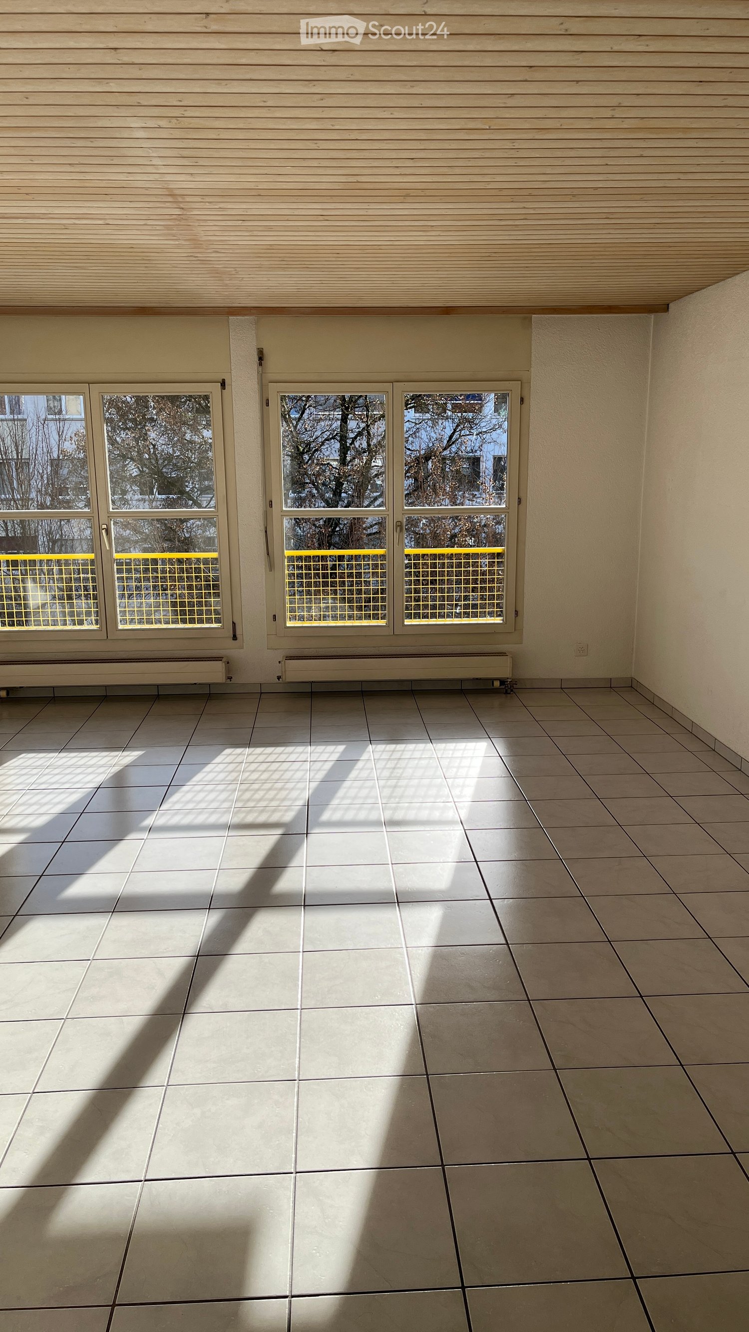 Empty room with white tile flooring, large windows with yellow fences, a white wall, and ceiling lights