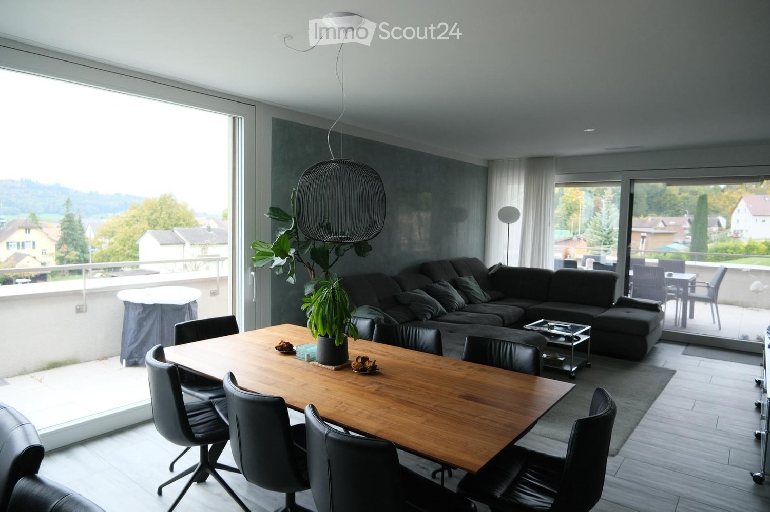 Large dining table, black leather chairs, gray rug, large windows, gray wall, plants, hanging lamp, couch on balcony