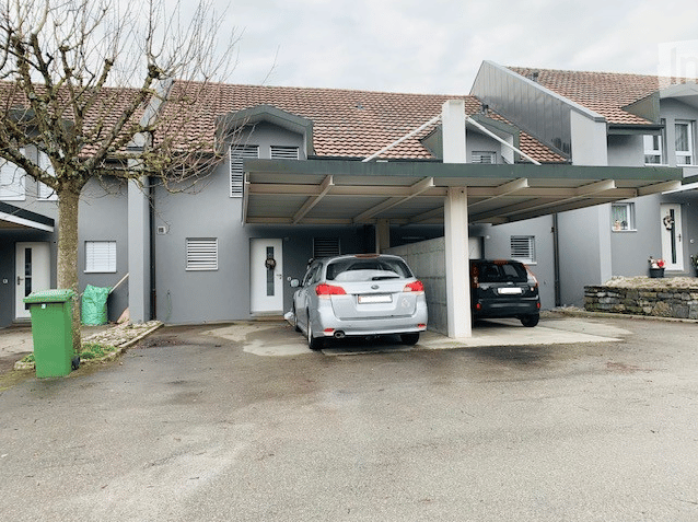 grey residential building with tiled roof, covered parking lot, two cars parked, two trash bins, tree, small stone wall