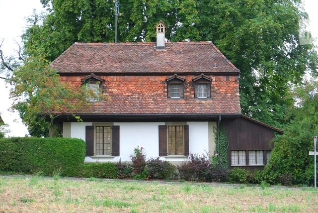 2 story house, brown roof, white walls, large chimney, multiple windows, surrounded by greenery and grass