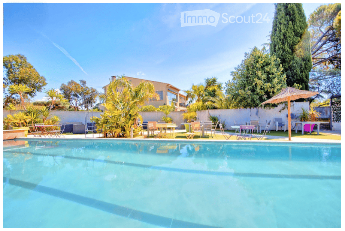 Outdoor area with a large swimming pool, surrounded by trees and tables and chairs, and umbrellas.