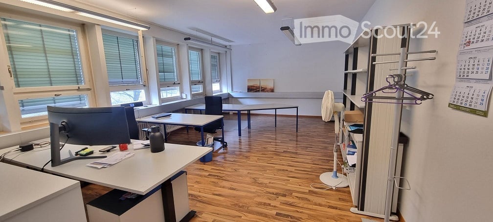 An office room with wooden floors, several desks and chairs, a monitor, office items, a cabinet, a fan, and a calendar on the wall.