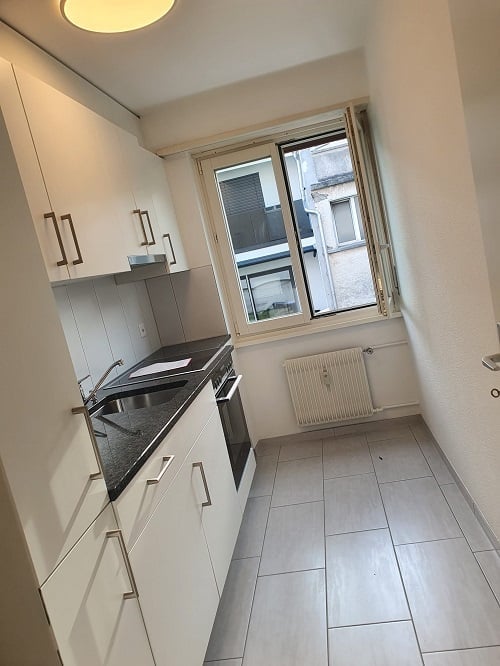 Kitchen area, tiled floor, white cabinets, black countertop, stainless steel sink, oven, white fridge, radiator, window