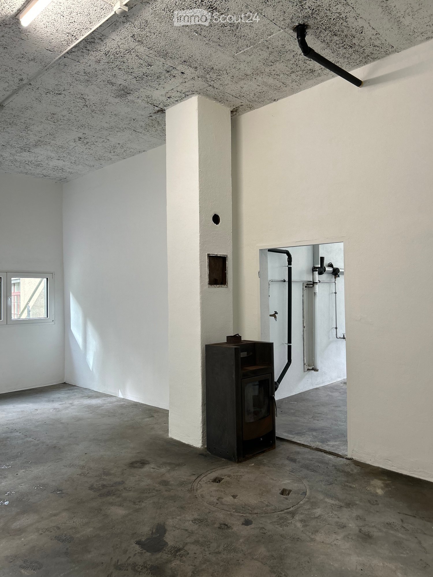Empty room with white walls, concrete floor, and a small window. A black stove is placed against one wall, and there is a small door leading to another area with visible pipes.