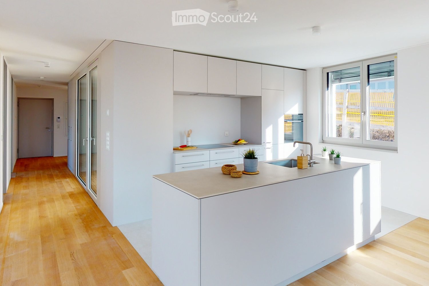 Modern kitchen with white cabinetry, wooden floors, island countertop, sink, faucet, potted plants, kitchen utensils, large window, sliding glass door, hallway leading to another room.