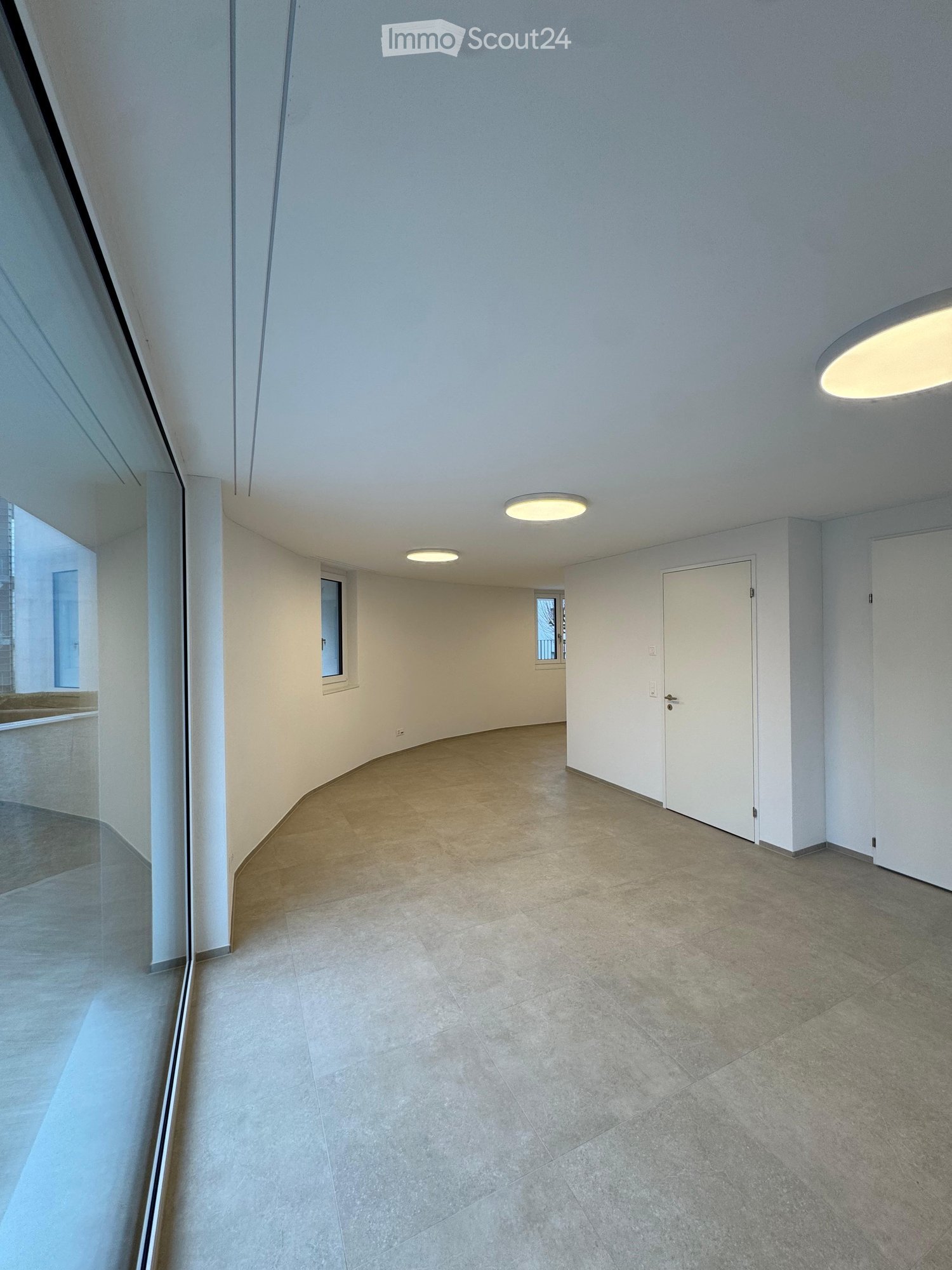 Large empty room with arched design, large glass windows, white tiled floor, and ceiling lights