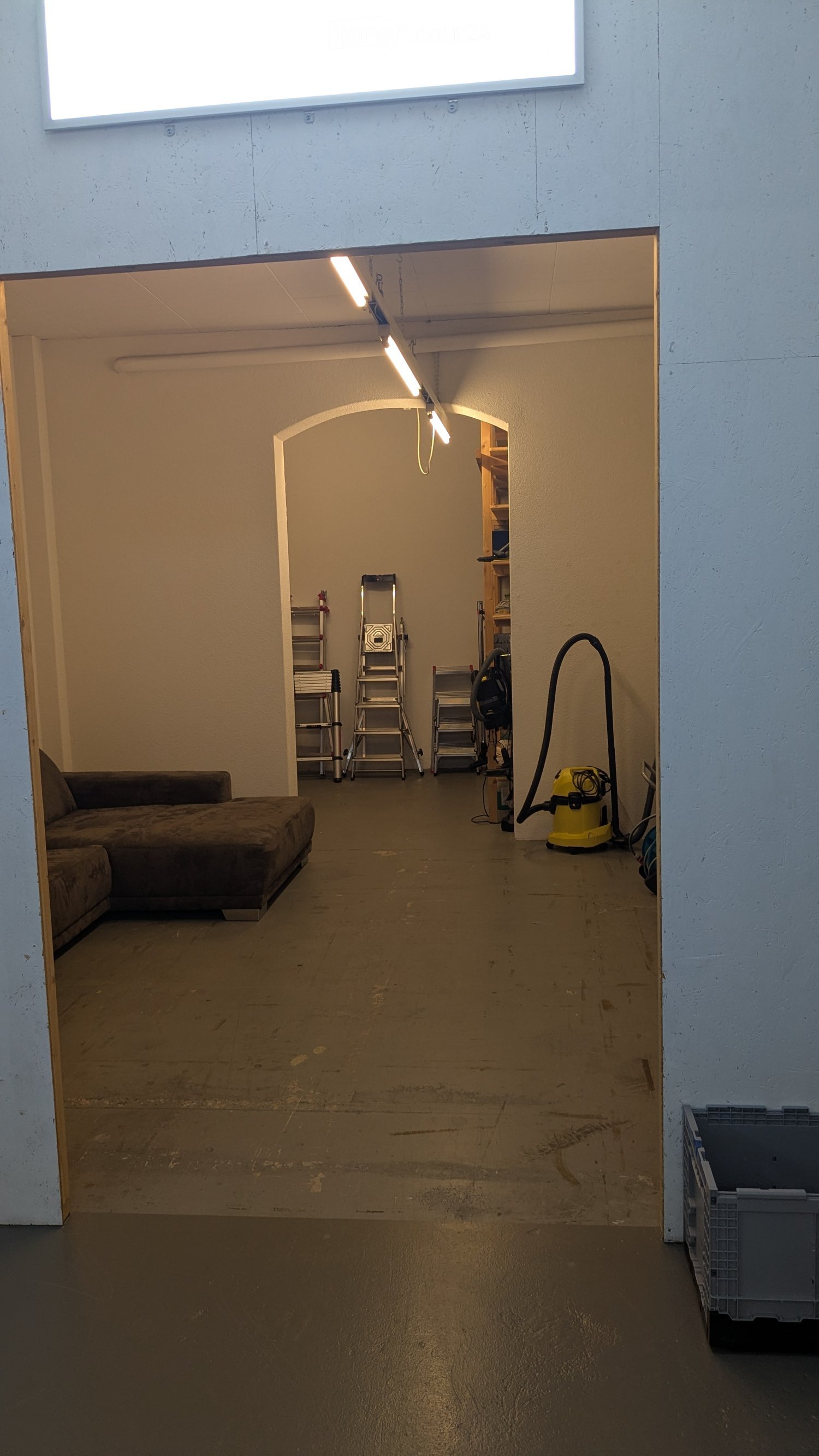 Empty storage room with light grey walls, concrete floor, sofa, ladders, storage box