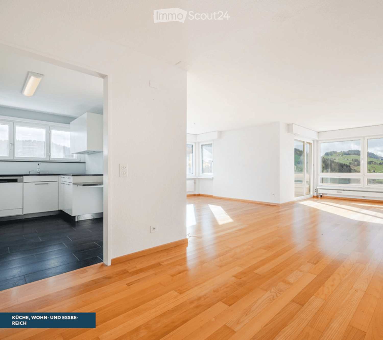 Open-plan space with wooden flooring, white walls, and large windows. The area includes a kitchen section with cabinets and a sink, and a living area with ample natural light.