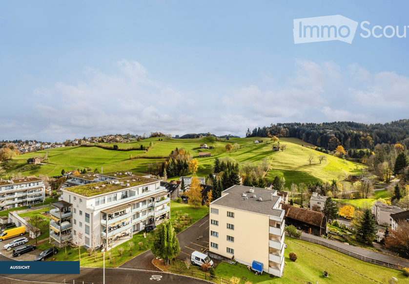 Aerial view of residential buildings, green fields, trees, and parked vehicles in a scenic landscape.