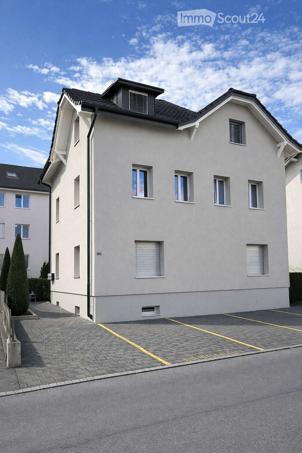 Two story house with beige walls and black roof, multiple windows, two parking places in front