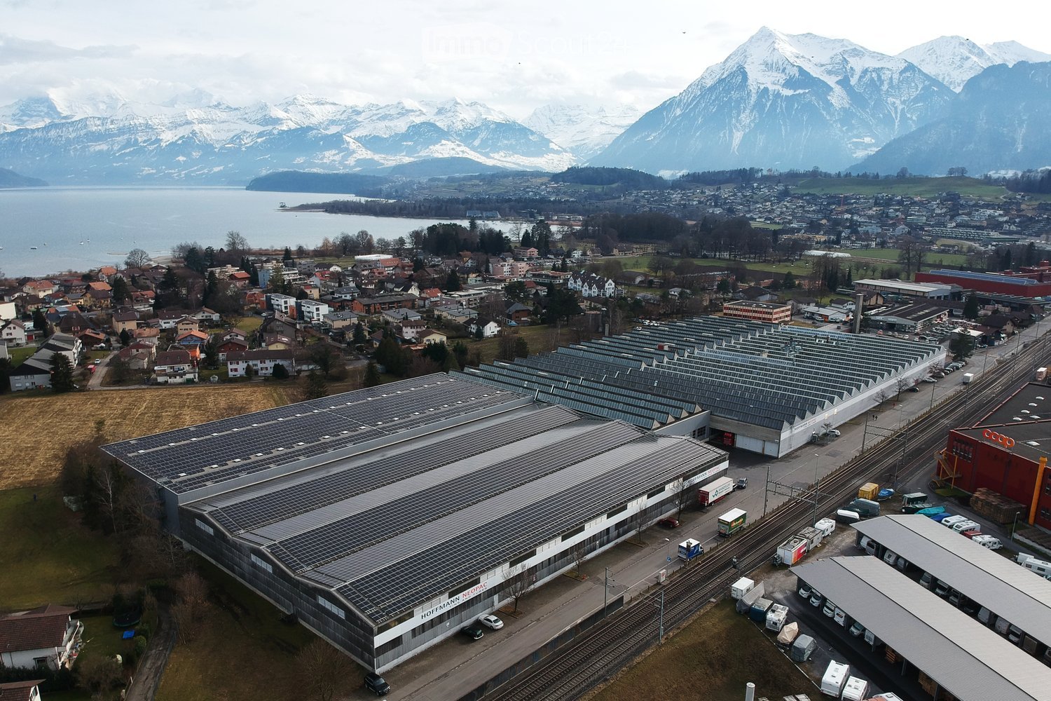 Aerial view of a large industrial building with solar panels on the roof, surrounded by smaller structures, parking lots, and a scenic backdrop of mountains and a lake.
