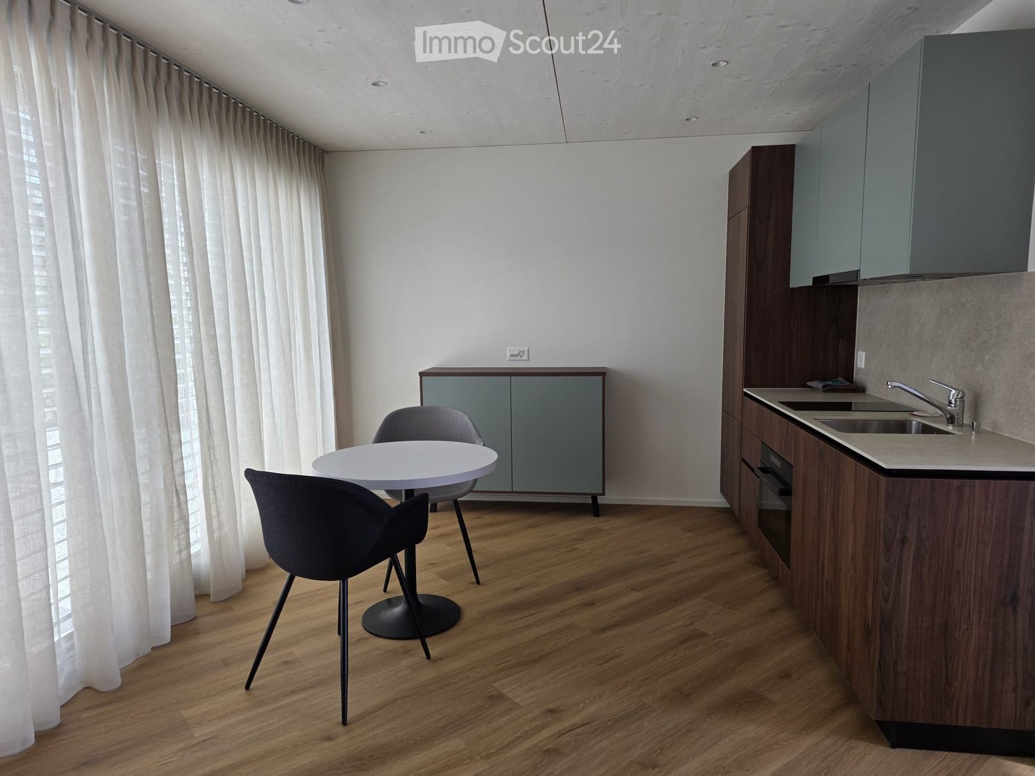 Modern kitchen with wooden floor, white walls, and gray cabinets. Includes a round white table, two chairs, and a sink with a faucet. A power outlet is mounted on the wall.