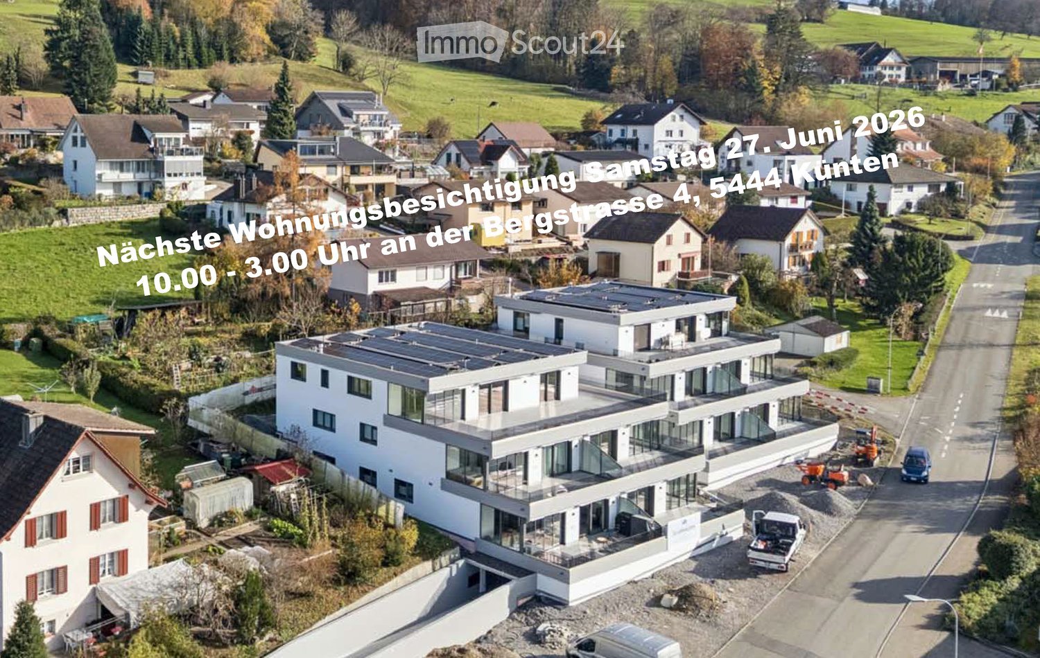 Aerial view of a modern two-story house with large windows and a flat roof, surrounded by a residential neighborhood with other houses and green fields.