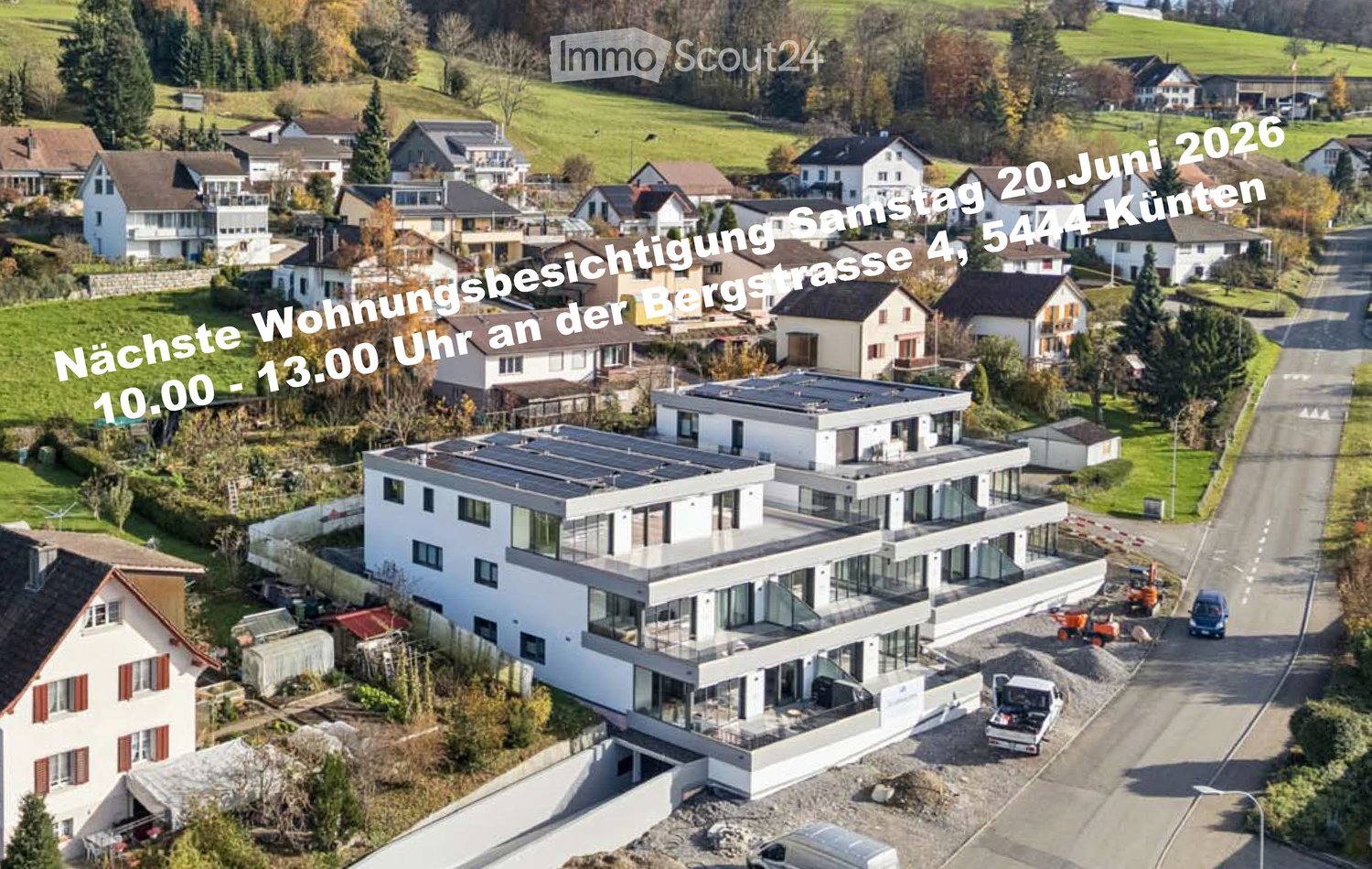 Aerial view of a modern two-story house with large windows and a flat roof, surrounded by other residential houses and green fields.