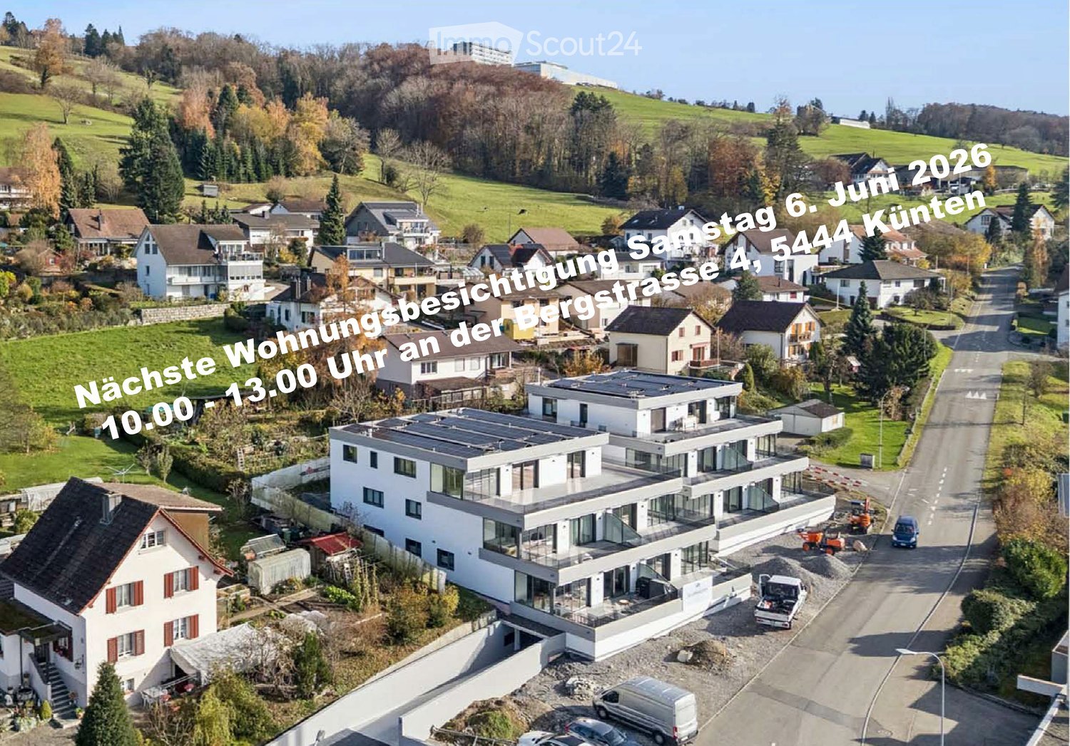 Aerial view of a modern two-story house with a flat roof and large windows, surrounded by a suburban neighborhood with other houses, green fields, and a road.