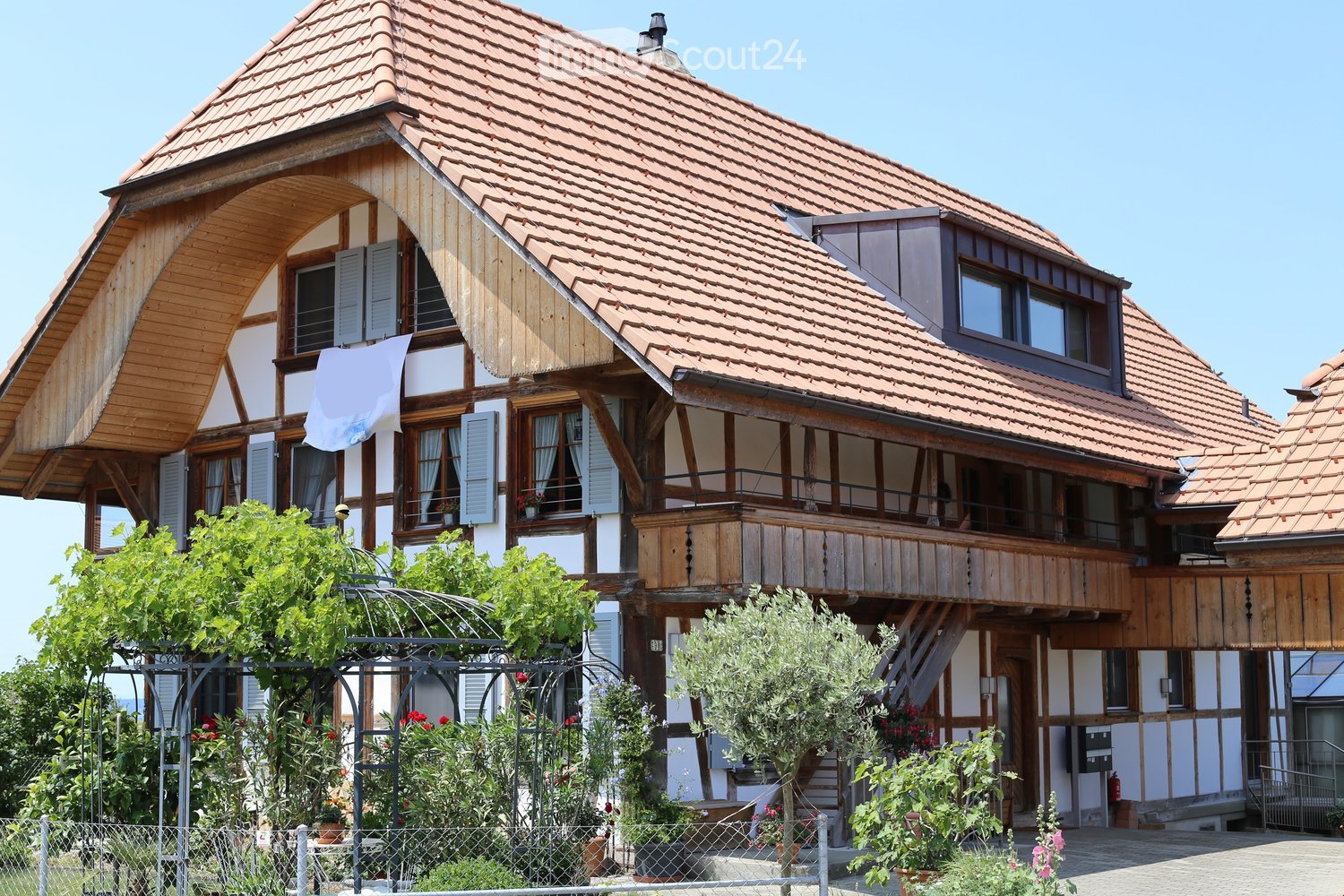 2-story house, wooden beams, red roof, white trim, windows with shutters, balcony with railings, garden with plants and gazebo