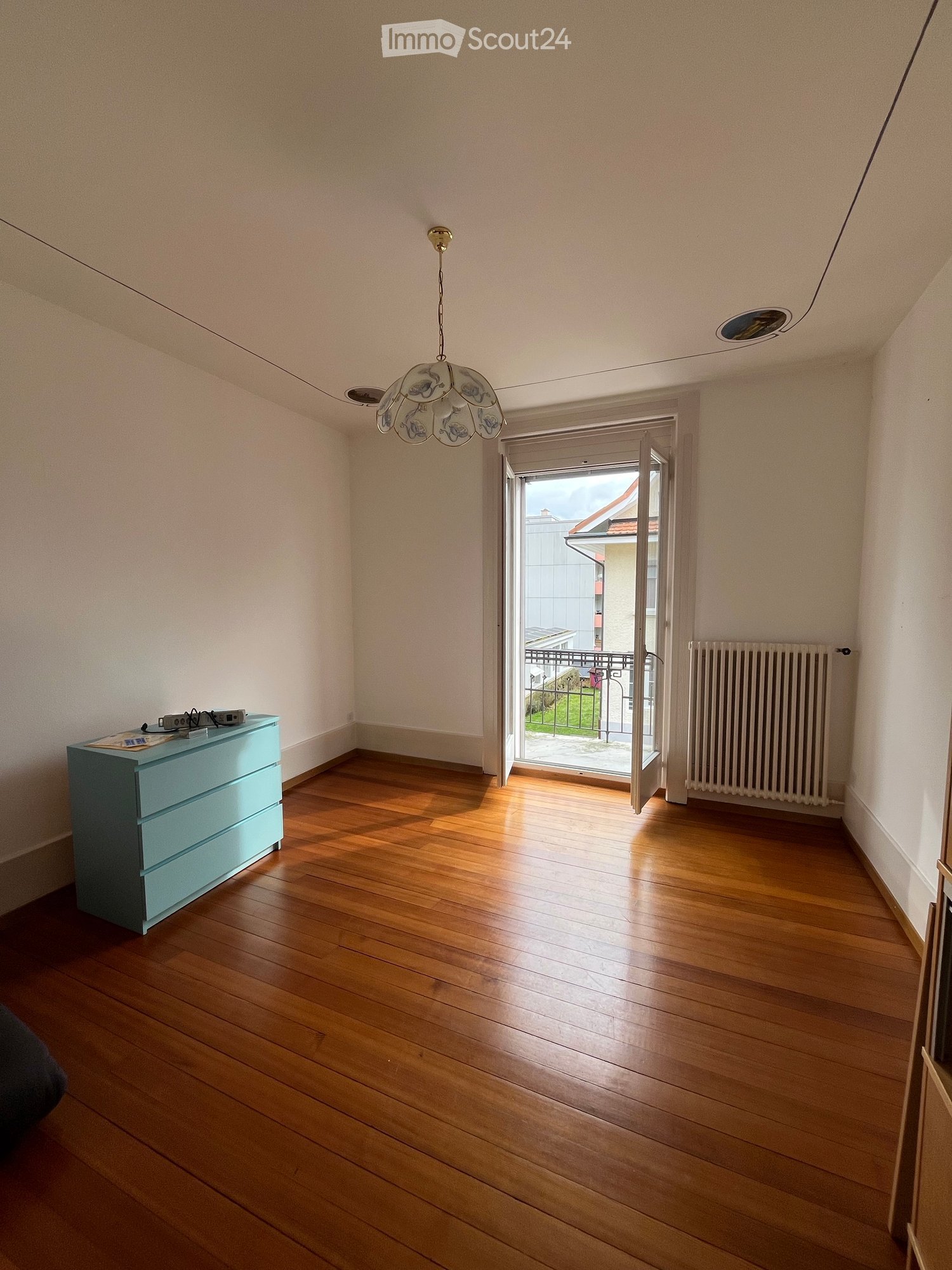 empty room, wooden floor, white walls, large windows, open door to balcony, radiator, light fixture