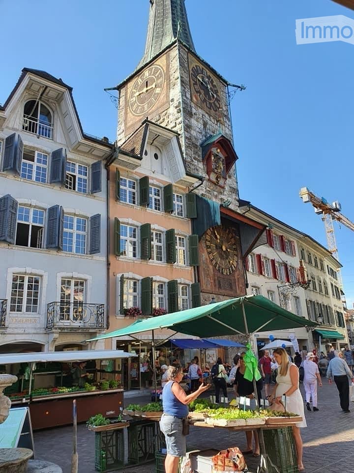 Town square with buildings, large clock tower, green market umbrellas, plants for sale